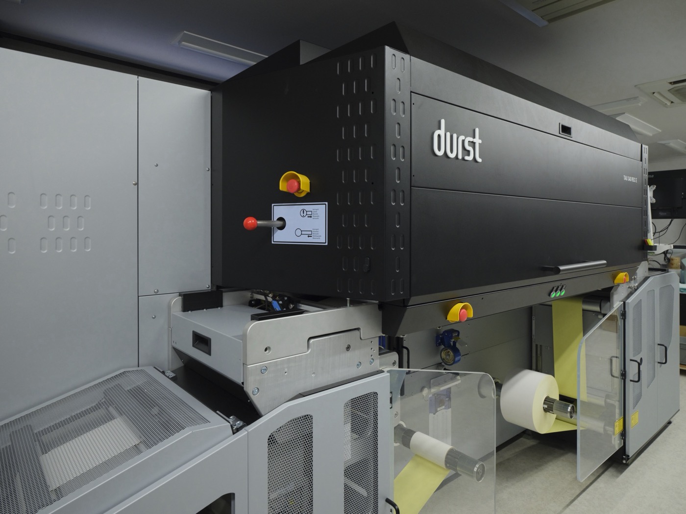 デジタル印刷機 Durst Tau 340 RSC E の本体クローズアップ。黒い筐体にdurstのロゴと黄色い非常停止ボタン、下部に黄色いロール紙